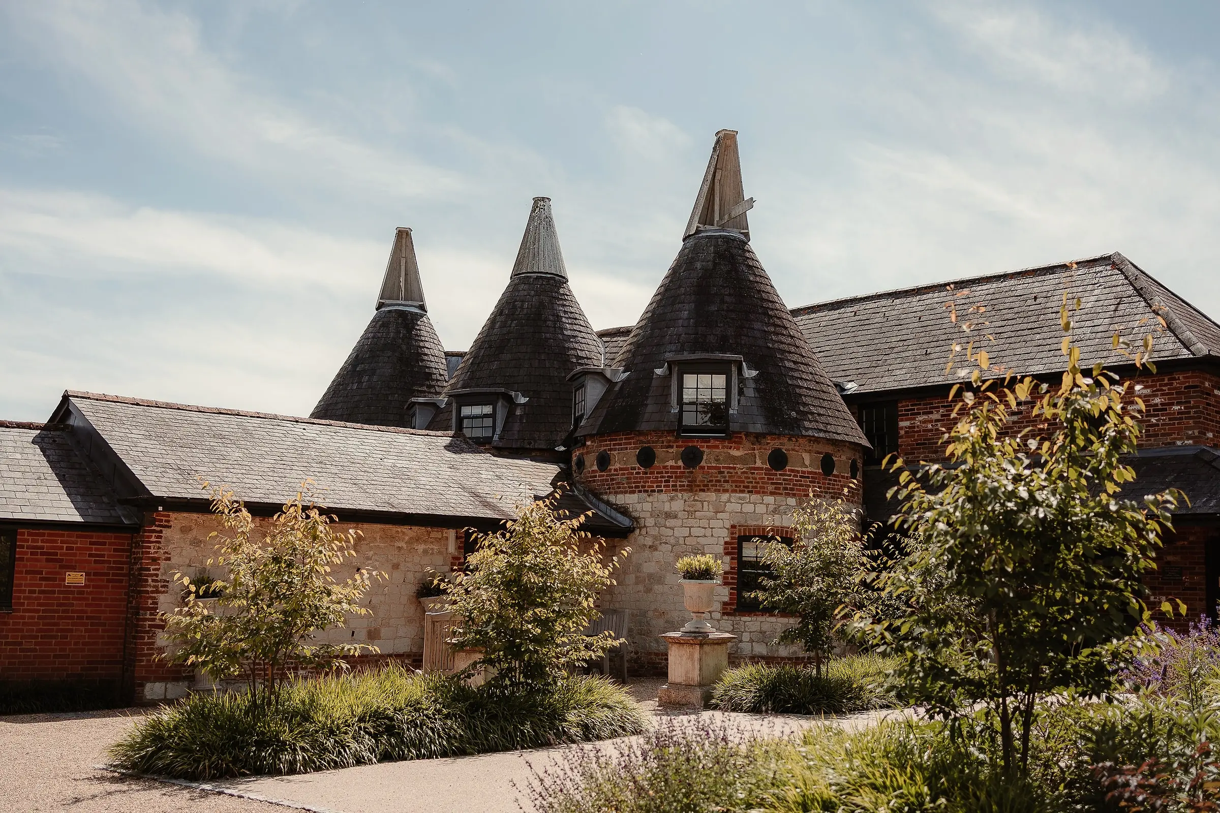 Three oast cowls above the red-brick and flint buildings, with young trees and grasses in front