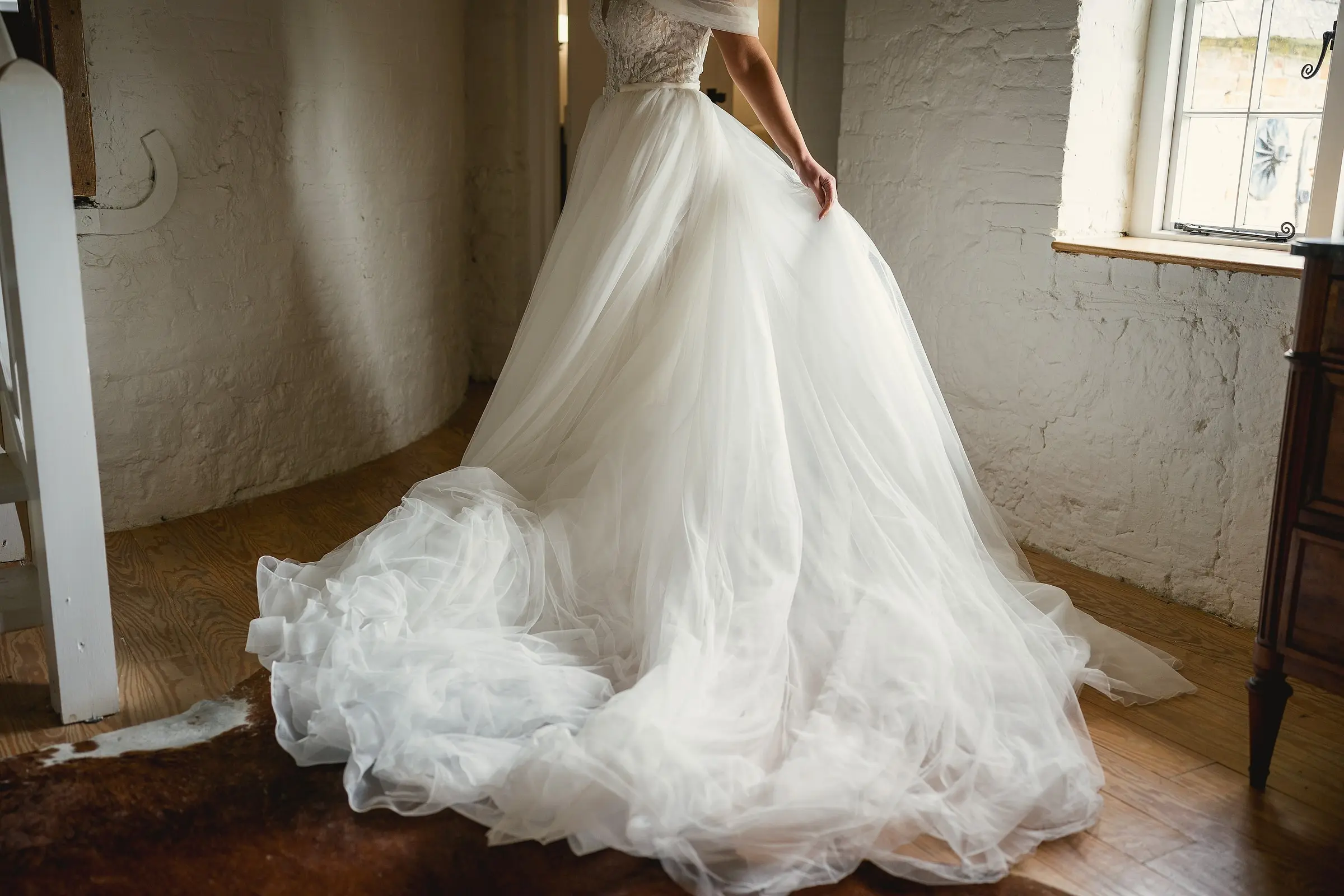 A bride's full tulle skirt spread across the floor of a white-walled room lit by a tall window