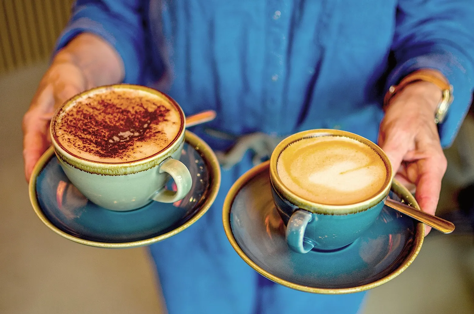 Two cappuccinos in teal cups carried out by a member of café staff