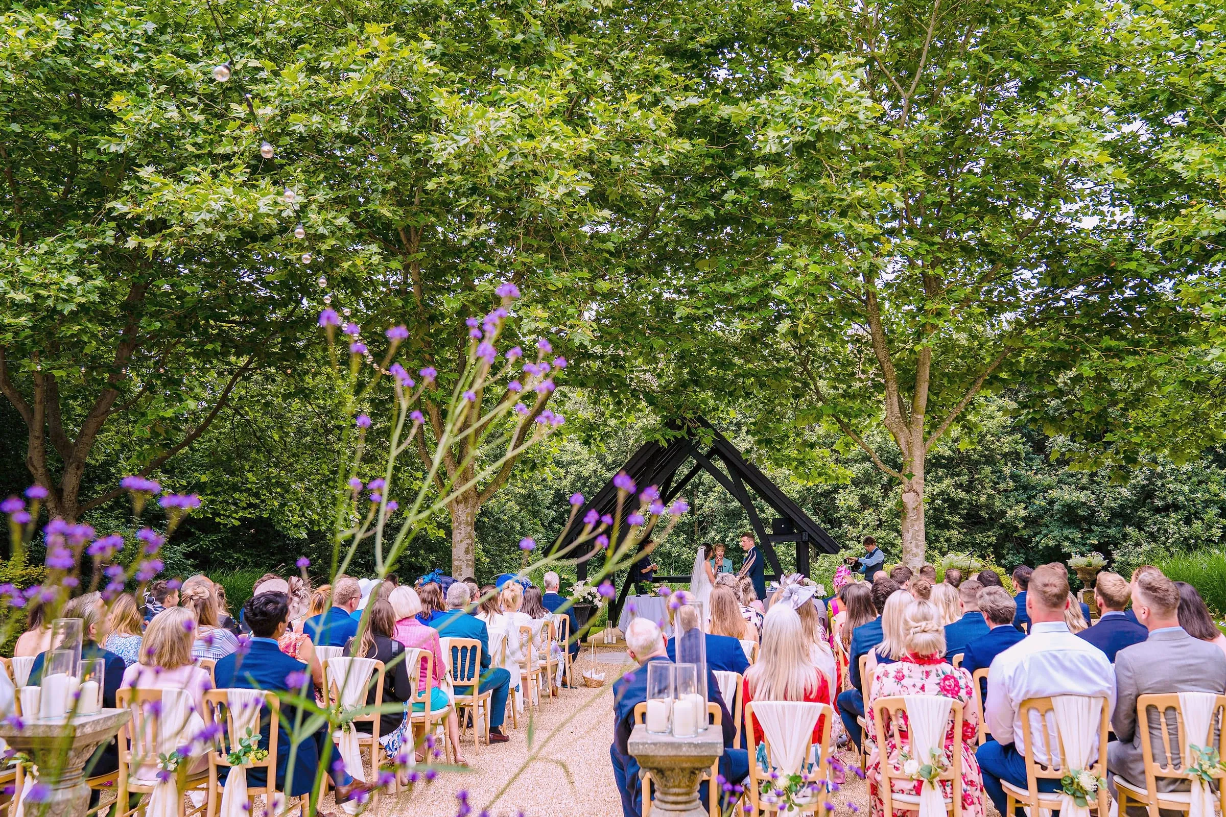 Guests watch an outdoor ceremony in the garden, verbena in the foreground and trees overhead