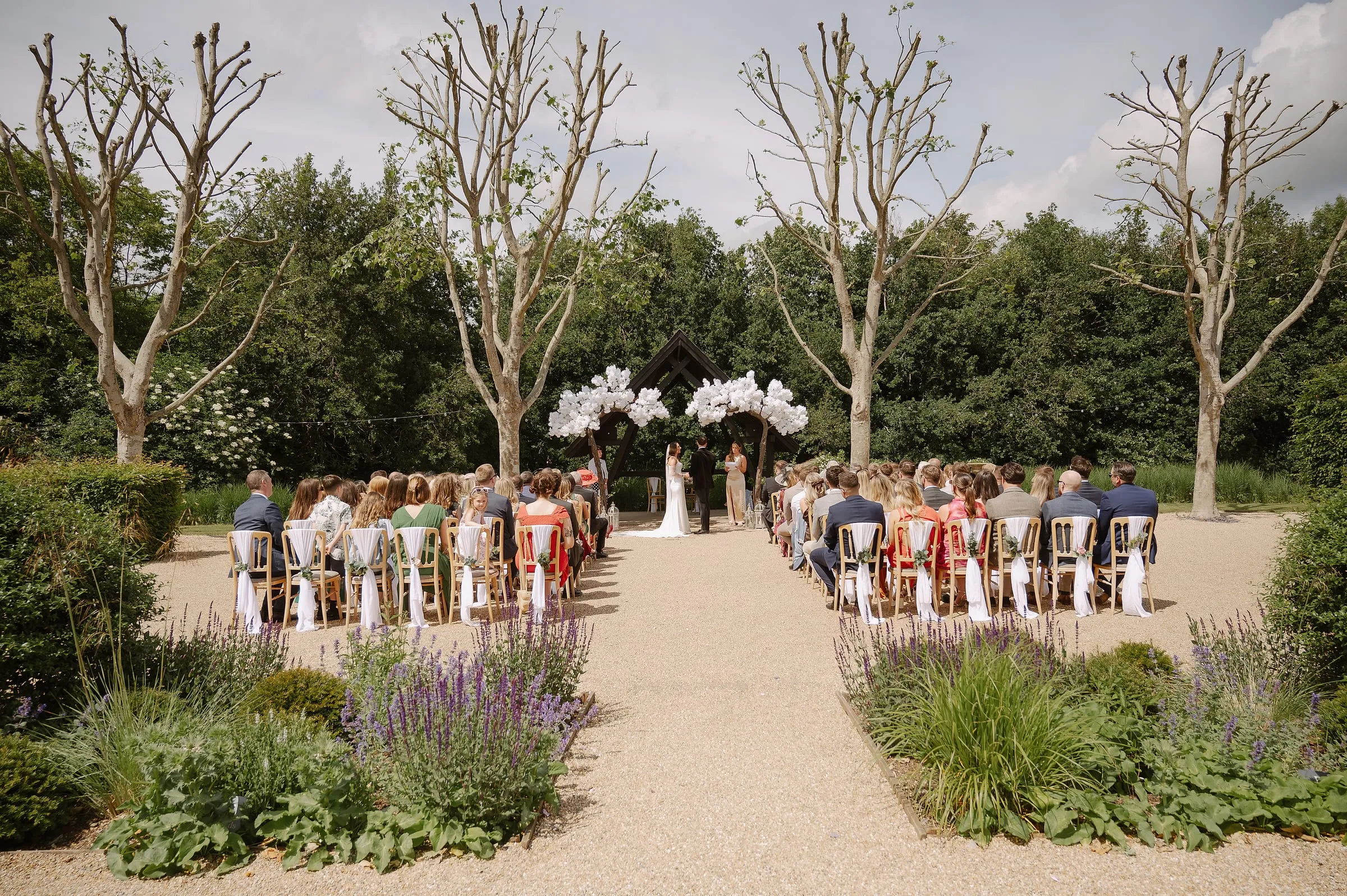 Guests seated either side of a gravel aisle as a couple marry under white blossom arches in the garden