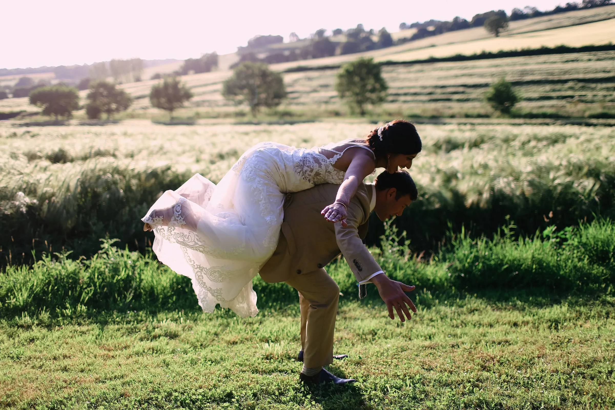 Groom carrying the bride piggyback across a sunlit meadow with rolling farmland behind