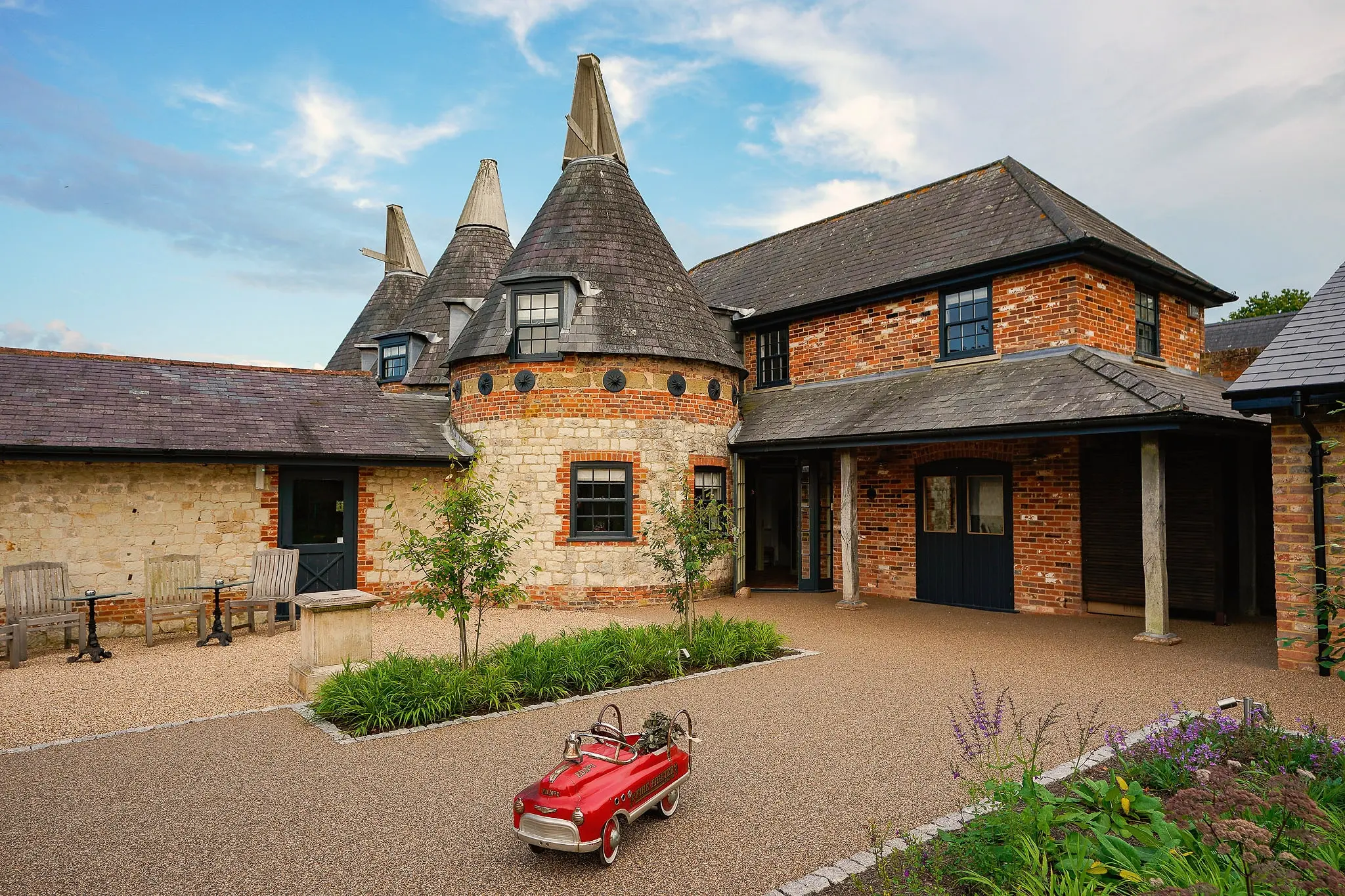 The oast house and red-brick barn around a gravel courtyard with planting and a toy red pedal car