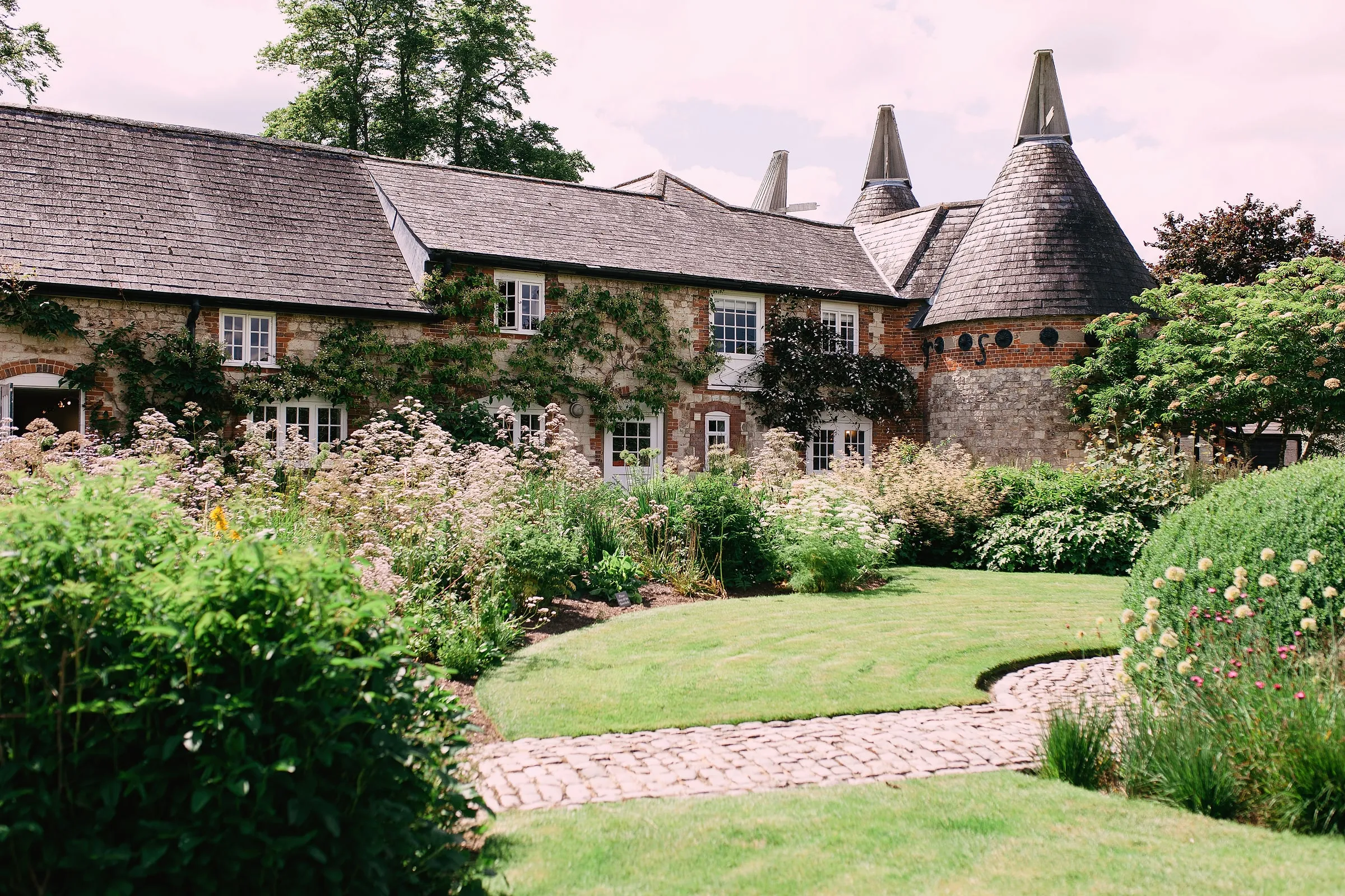 Curving lawn, cobbled path and summer borders in front of the flint house and oast roundel