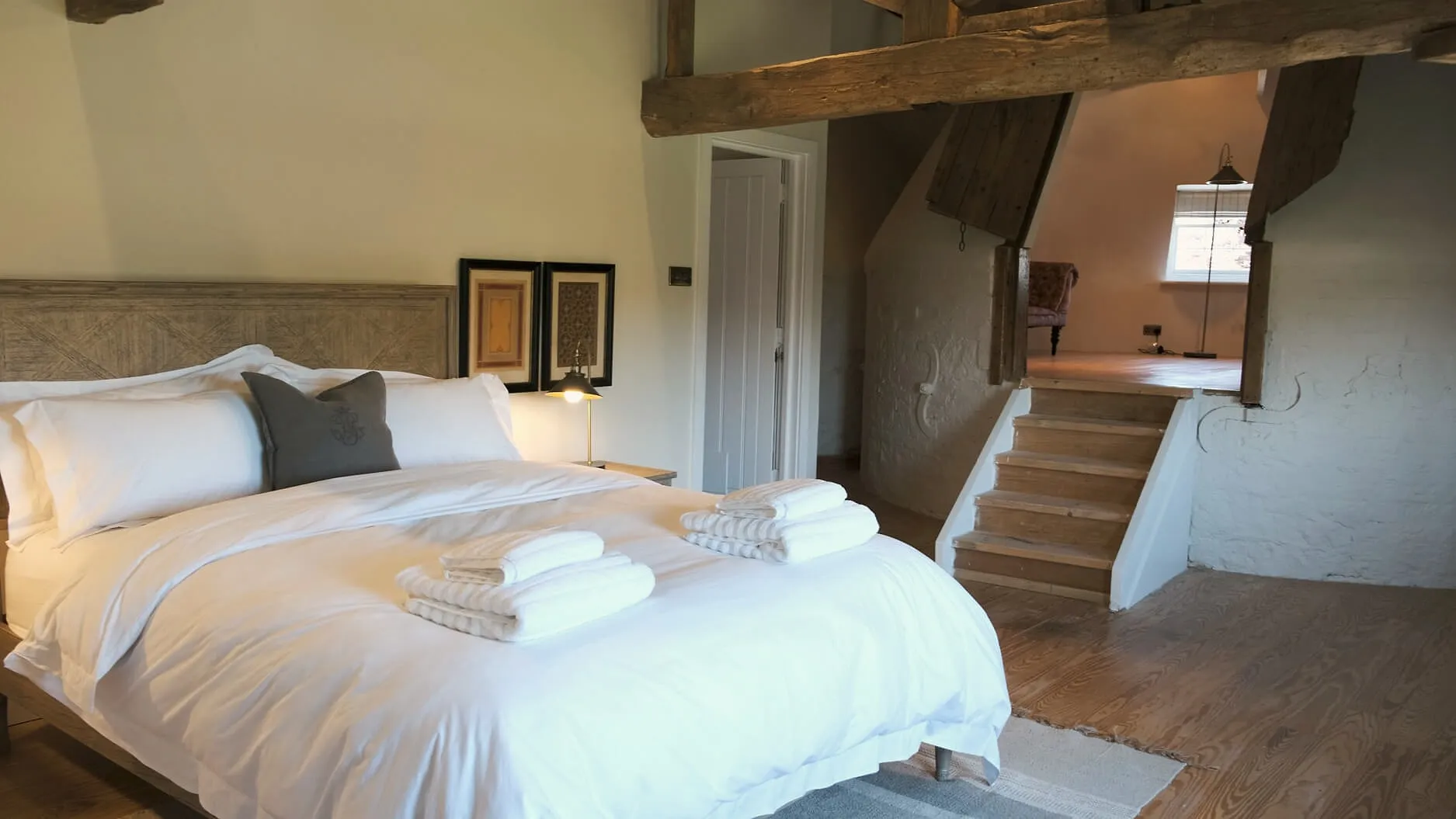 A double bed with folded white towels under an old oak beam, steps leading up to a landing