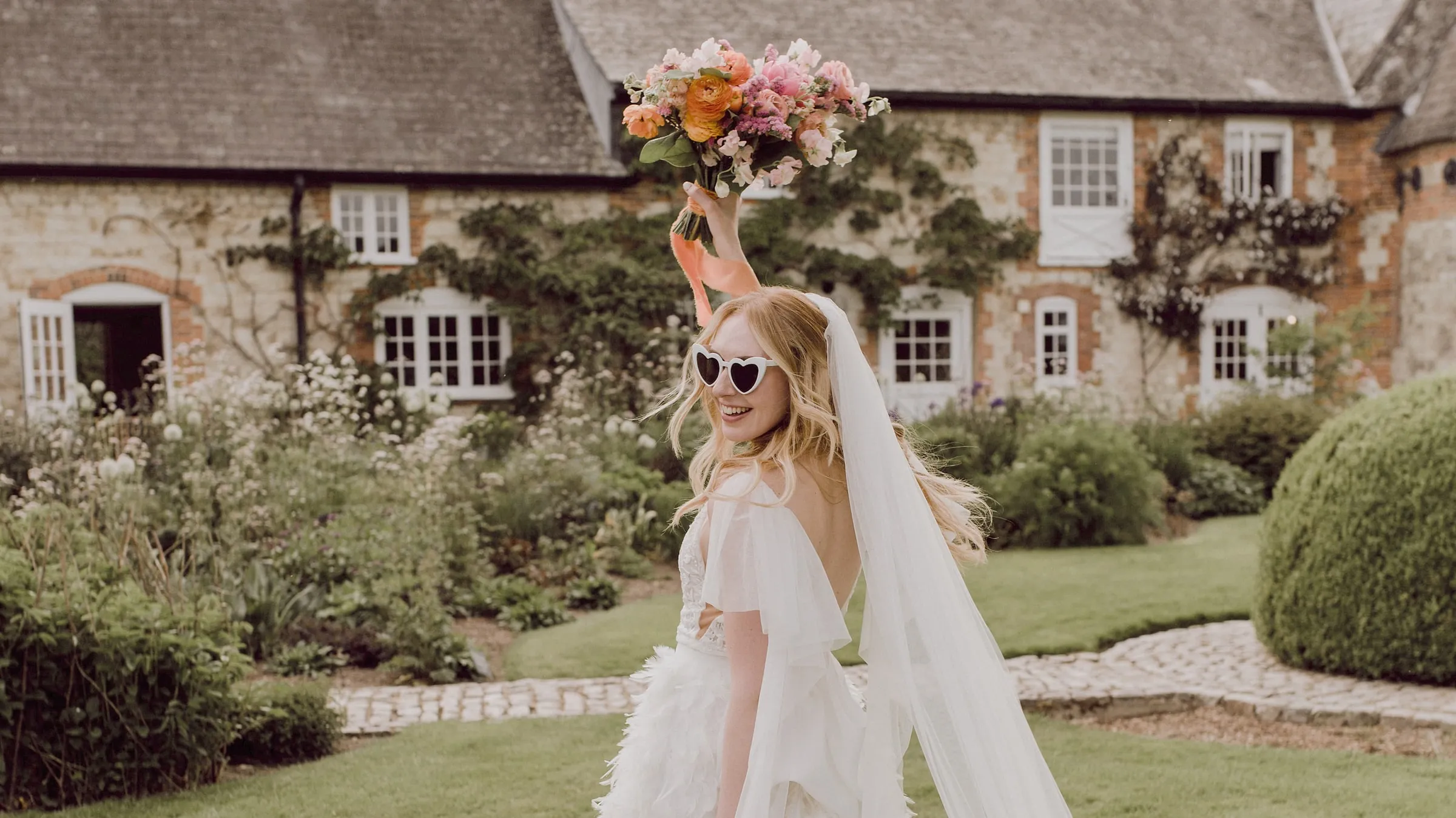 Bride in heart-shaped sunglasses holds her bouquet high on the lawn in front of the flint house